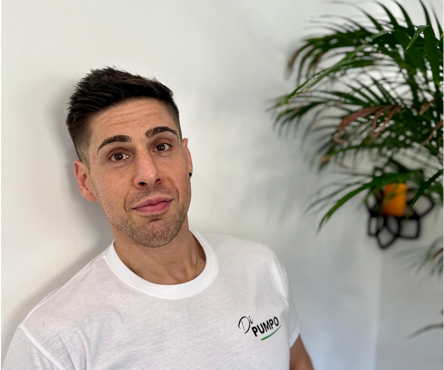 Junger Mann in weißem T-Shirt vor einer Wand mit Pflanze im Hintergrund.